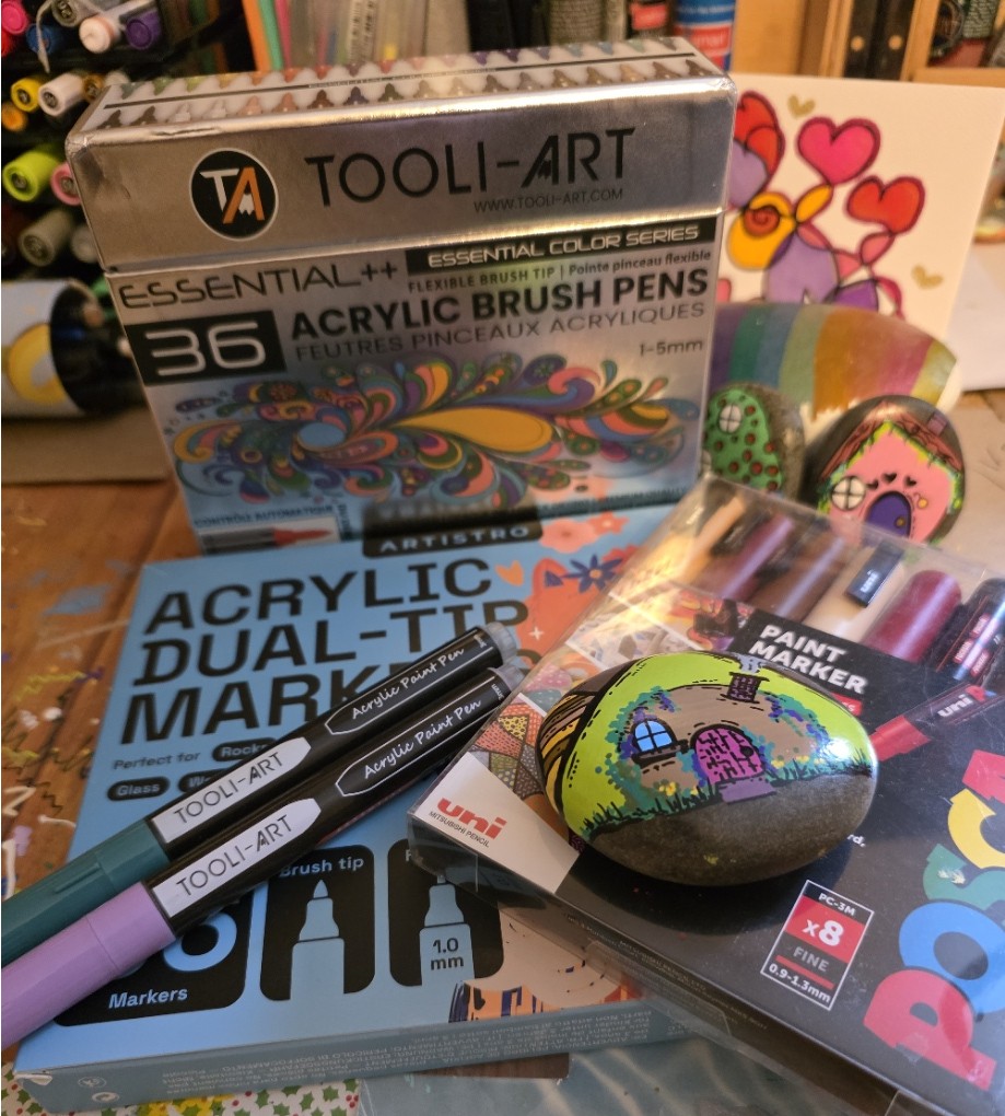 A display of three brands of acrylic paint markers used for painting kindness rocks. There are also three painted rocks in the picture. 
