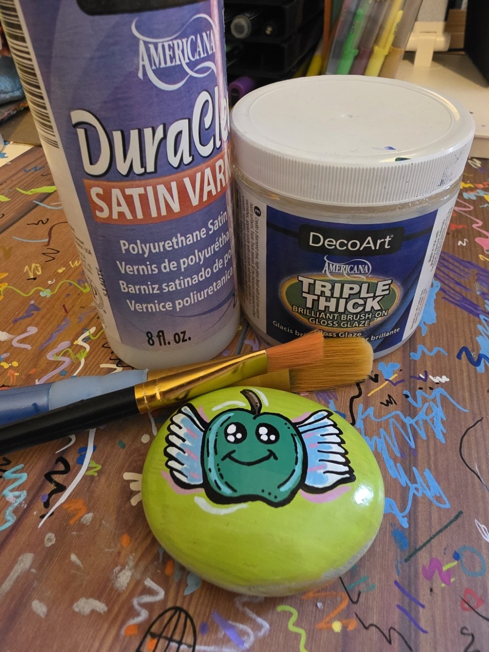 A picture of two outdoor sealers from the brand Duraclear, alongside paint brushes and a painted kindness rock. 