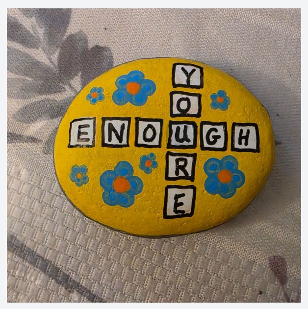 Bright yellow painted rock with a scrabble layout and the words "You Are Enough" with blue flowers around it. 